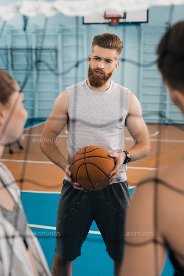 Bearded young sportsman with ball talking with teammates in sports hall