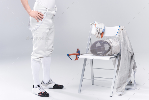 Low section of young man professional fencer standing near chair with fencing equipment isolated on
