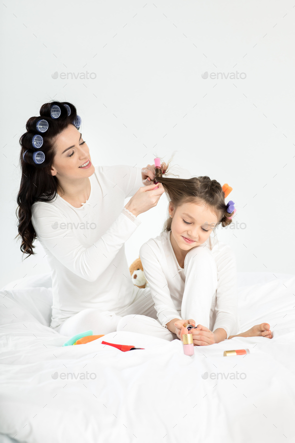 Mother curling hair to daughter applying nail polish on toenails isolated on white