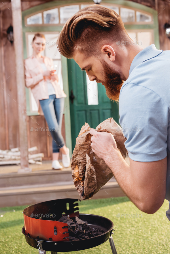 Young woman standing on porch and looking at bearded man holding coal for outdoor grill