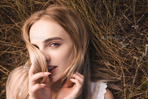 top view of sensual blonde woman lying on green grass, obscuring face with hair and looking at