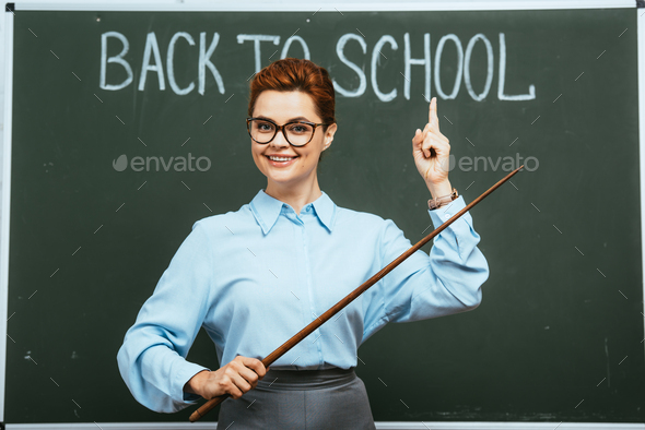 smiling teacher holding pointer and pointing with finger at back to school inscription on chalkboard
