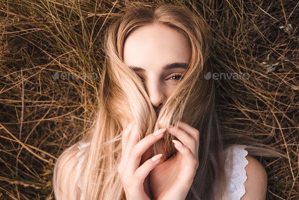 top view of young blonde woman obscuring face with hair while lying on grass and looking at camera