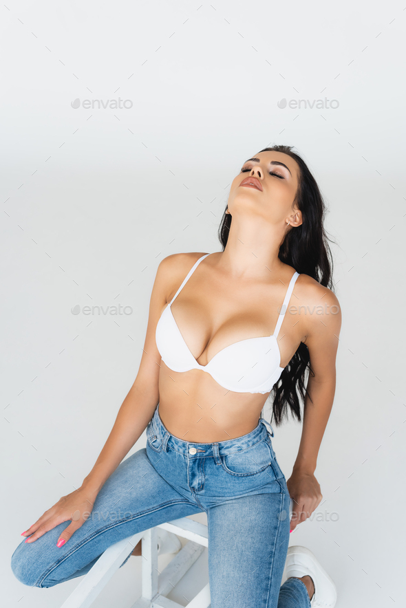 sexy woman with closed eyes in jeans and bra sitting on chair isolated on  grey Stock Photo by LightFieldStudios