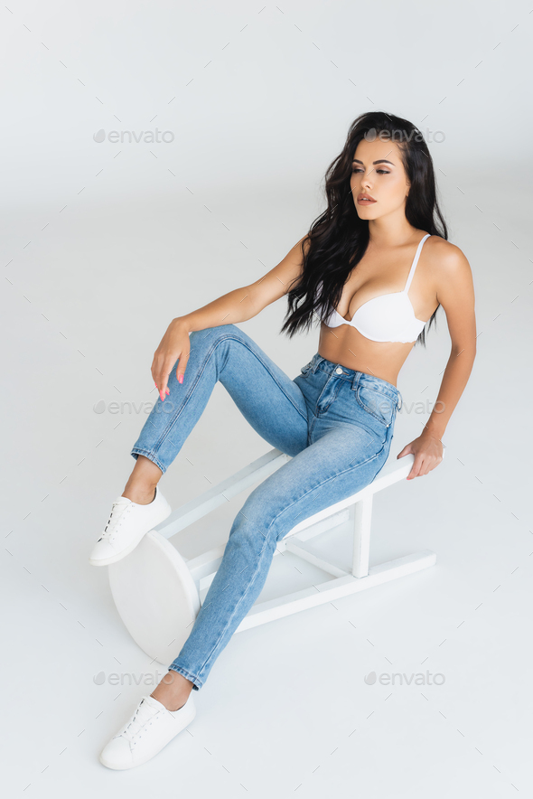 brunette woman in jeans and bra sitting on stool on grey Stock