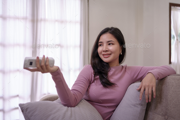 Young Asian woman sitting indoor at home watch tv holding remote control on sofa.