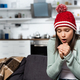cold woman in warm hat blowing on clenched hands while sitting on sofa in kitchen - PhotoDune Item for Sale