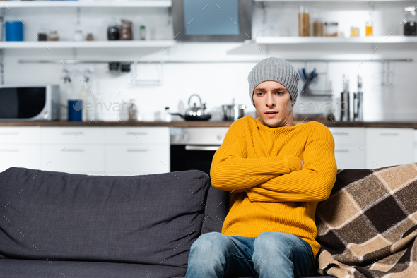 shivering man in knitted sweater and hat sitting on sofa with crossed arms in cold kitchen - Stock Photo - Images