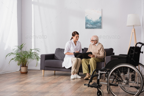 geriatric nurse and elderly man browsing photo album on blurred foreground - Stock Photo - Images