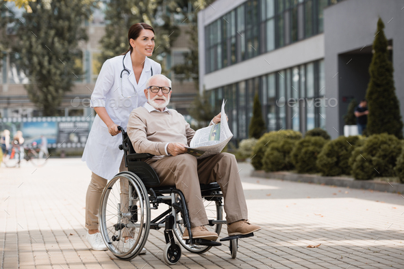 geriatric nurse walking with elderly disabled man in wheelchair with newspaper outdoors - Stock Photo - Images