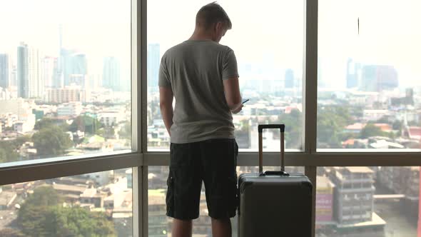 Young Man Uses Mobile Phone with Luggage Waits in the Station with Big Windows City View