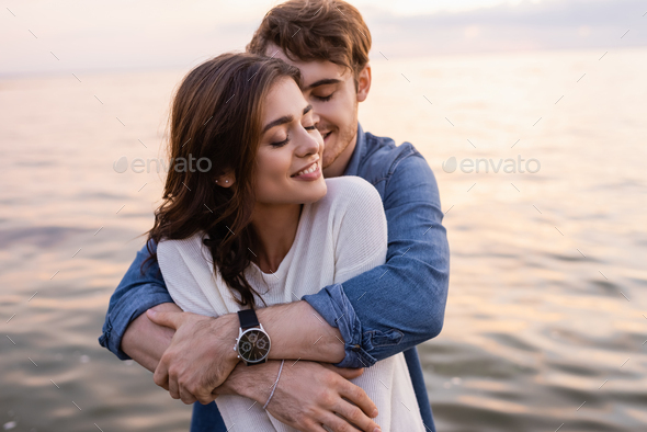 Young man hugging brunette girlfriend with closed eyes beside sea at sunset - Stock Photo - Images