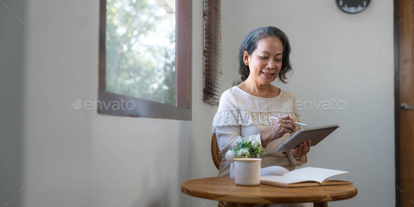60s aged Asian woman in casual clothes sitting by the window in her living room and using portable - Stock Photo - Images