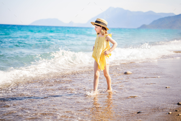 Little girl walking on beach, sea ocean shore in romantic yellow dress, straw hat. Playing in  - Stock Photo - Images