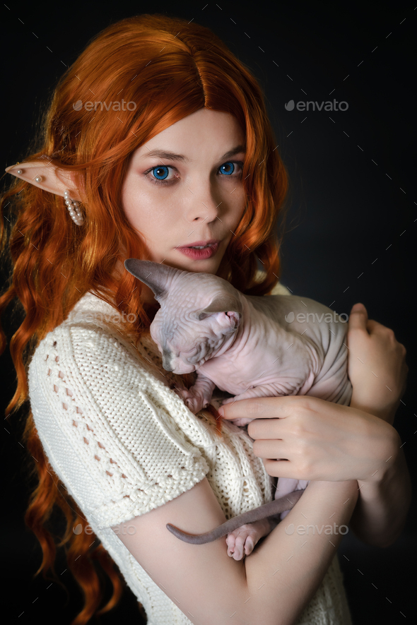 Portrait of young woman cosplayer elf in white knitted dress hugging Sphinx kitten. Black background - Stock Photo - Images