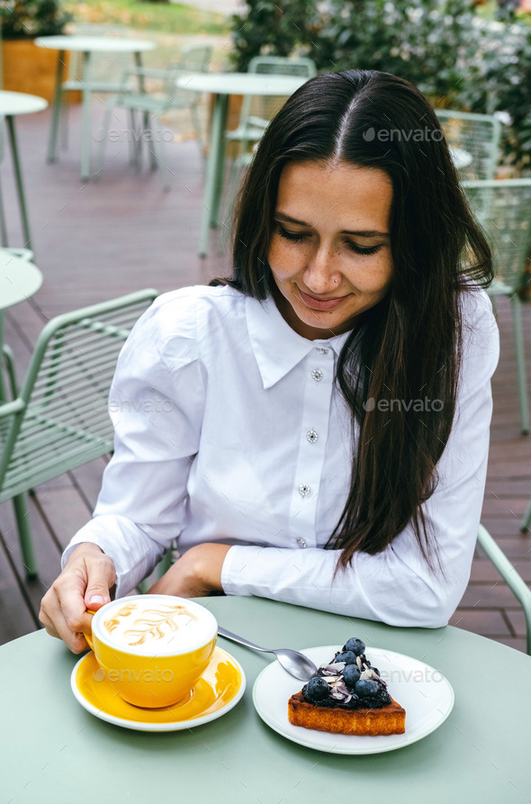 Happy brunette woman drinking coffee and eating blueberry cheesecake at outdoor cafe.  - Stock Photo - Images