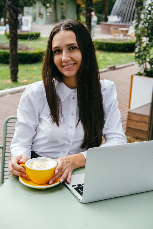 Young woman works at laptop and drinks coffee on the terrace in cafe. Lifestyle portrait. - Stock Photo - Images