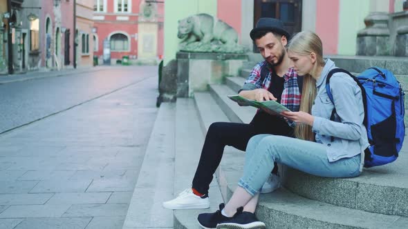 Tourist Couple Sitting on Street Stairs Checking City Map and Discussing alt