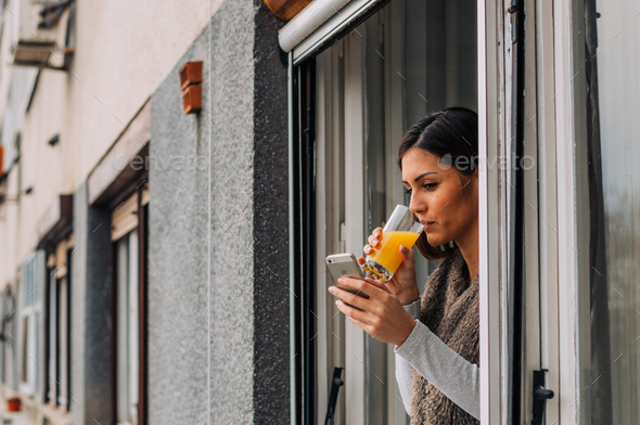 A girl standing by the window, using a phone, drinking orange juice. - Stock Photo - Images