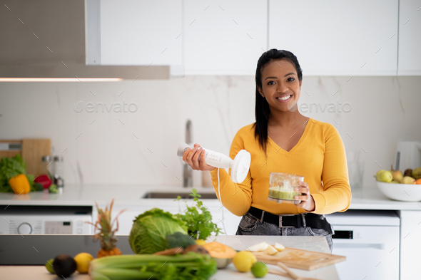 Smiling millennial african american lady in casual making smoothie with blender enjoy health care