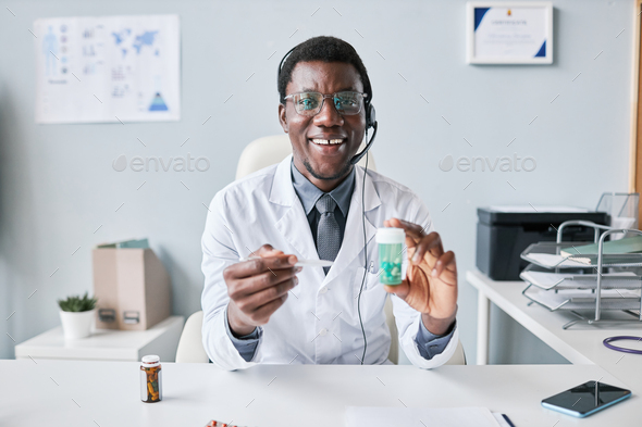 Black doctor holding bottle of pills wearing headset at workplace in office - Stock Photo - Images