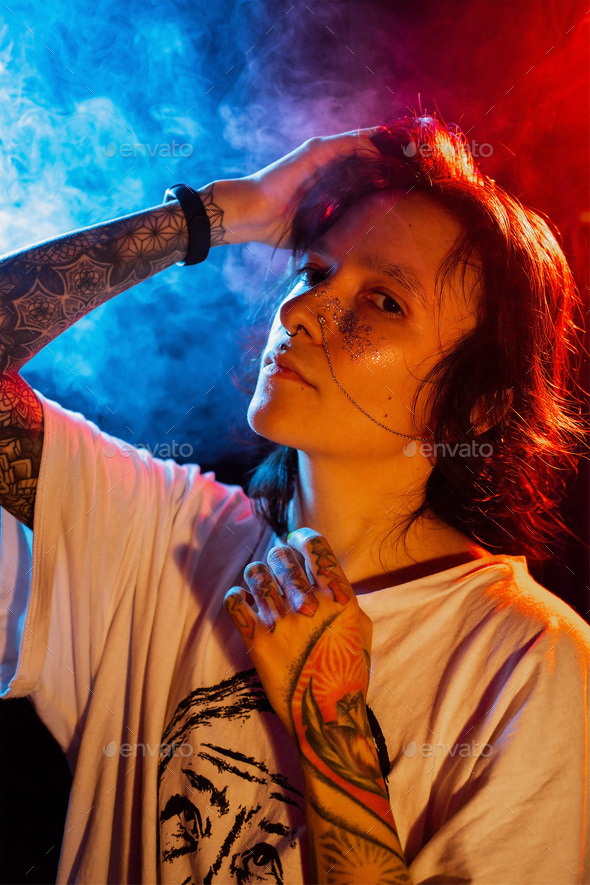 Stylish young tattooed woman with face piercing posing on dark background with smoke in neon lights. - Stock Photo - Images