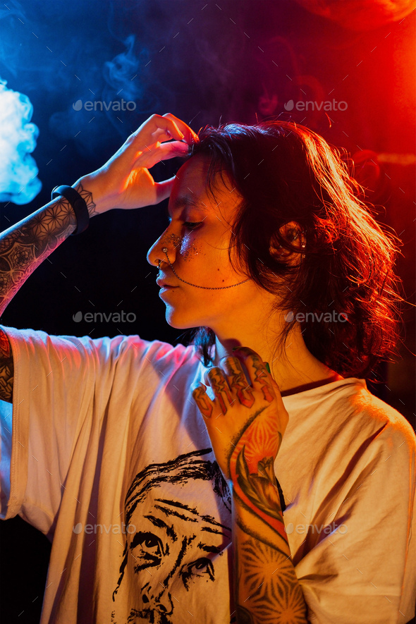 Stylish young tattooed woman with face piercing posing on dark background with smoke in neon lights. - Stock Photo - Images