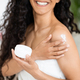 Cheerful islamic millennial curly lady in towel holding jar with copy space, apply cream on shoulder - PhotoDune Item for Sale