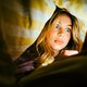 Young amazed caucasian woman using smartphone, lies under a blanket on a bed at home in the evening. - PhotoDune Item for Sale