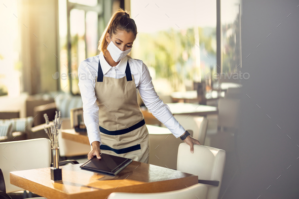 Waitress wearing protective face mask while preparing tables for the guests in a restaurant.
