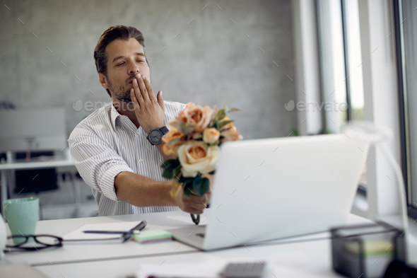 Affectionate businessman with bouquet of flowers sending a kiss while dating online in the office.