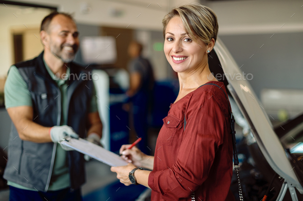 Happy woman signing paperwork at auto repair shop and looking at camera.