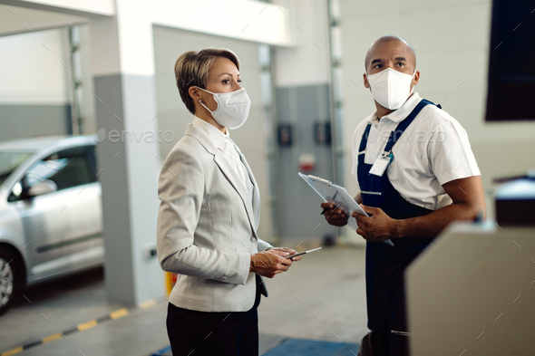 Black mechanic and businesswoman wearing face masks at auto repair shop due to COVID-19 pandemic.