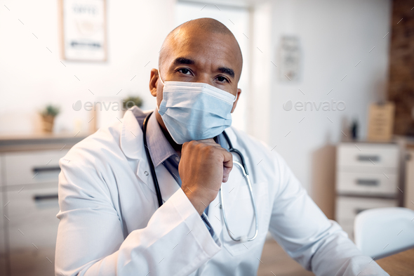 Male black doctor with protective face mask working at the clinic and looking at camera.