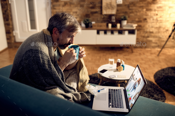 Mid adult man drinking tea while having video call over laptop with his doctor at night.