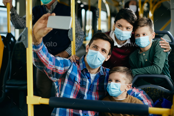 Happy family with face masks taking selfie while commuting by bus.