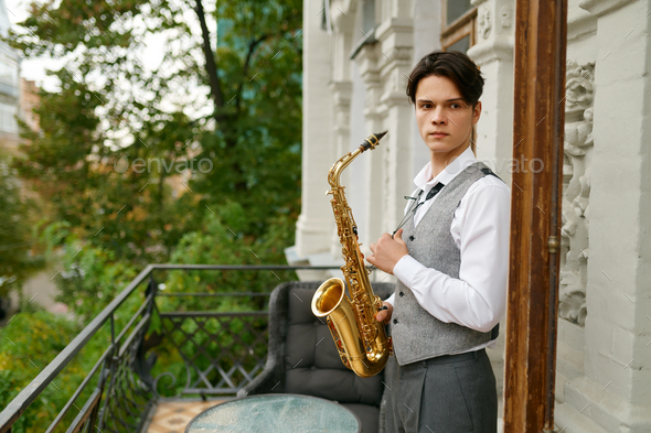 Young man holding saxophone standing on balcony terrace - Stock Photo - Images