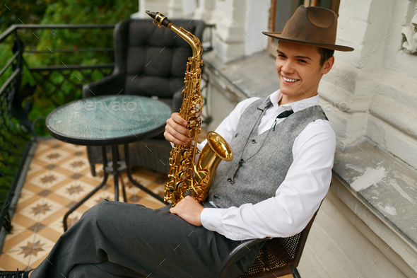 Smiling musician holding saxophone sitting on chair in balcony of his apartment - Stock Photo - Images