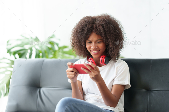 Beautiful young woman playing game on her mobile phone in her living room on sofa at home happily  - Stock Photo - Images