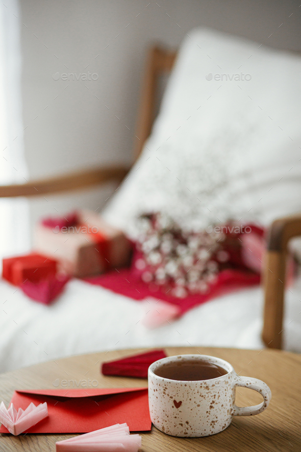 Cute stylish cup of tea with heart and red envelope on wooden table against modern armchair - Stock Photo - Images