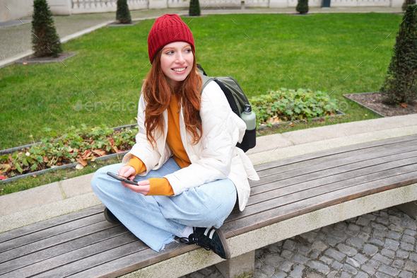 Portrait of stylish young woman, 25 years, sits on bench in park and uses mobile phone, reads