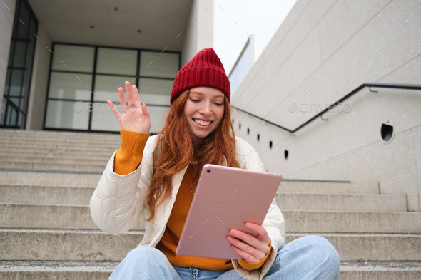 Joyful redhead teen girl, student with digital tablet, says hello, waves hand at gadget camera