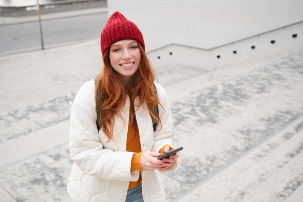 Smiling redhead girl, student tourist walks around city, goes up the stairs, looks at mobile phone