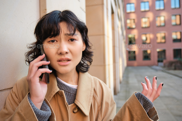 Korean girl with concerned face talks on mobile phone and shrugs, frowns and looks concerned, being