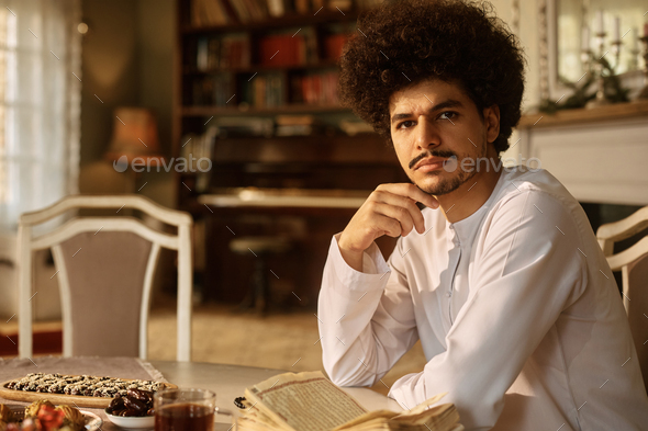 Intuïtie Bemiddelaar Bevestigen Young Arab man reading from Quran at dining table and looking at camera.  Stock Photo by drazenphoto