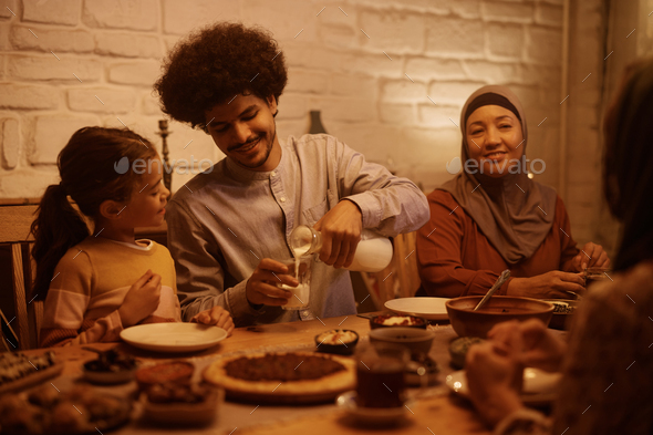 Happy Muslim father pouring milk into daughter's glass during family dinner at dinning table. - Stock Photo - Images