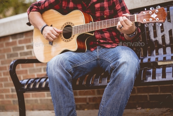 Male wearing a red and black flannel sitting on a bench playing the guitar