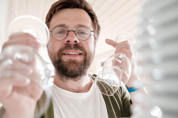 Camera view from the kitchen cabinet of a cute smiling man putting away clean wine glasses on shelf - Stock Photo - Images