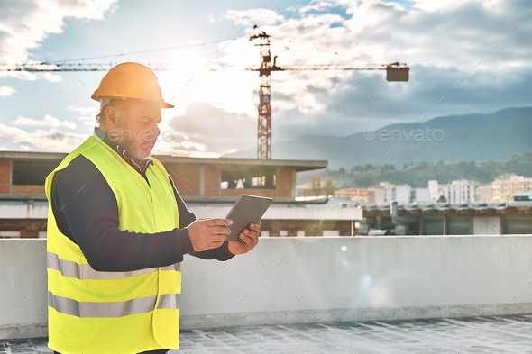 Middle aged bearded supervisor in hardhat and safety vest making notes in tablet on building site. - Stock Photo - Images