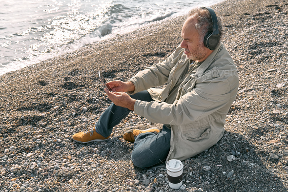 Bearded man listening music in headphones and drinking coffee while sitting on the beach. - Stock Photo - Images